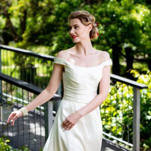 A woman with an elegant updo hairstyle featuring twists and a braided crown, complemented by tendrils framing the face, set against a natural outdoor backdrop.