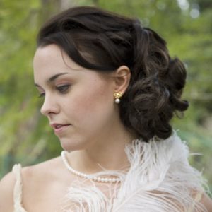 Woman with soft curls loosely gathered in side-swept updo at nape, complemented by vintage pearl earring and feathered garment