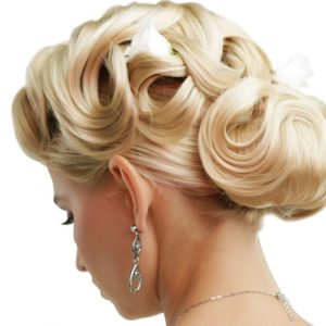 Profile view of elaborate blonde updo featuring large smooth curls and rolls adorned with small white flowers. Dangling earring and subtle necklace visible, plain white background