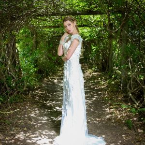 Woman in elegant white bridal gown with classic updo hairstyle featuring neatly twisted bun at back of head, posing in lush green wooded pathway