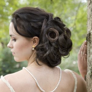 Brunette woman with soft, voluminous curled updo, strands pinned at back. Wearing pearl earrings and necklace, standing beside a tree